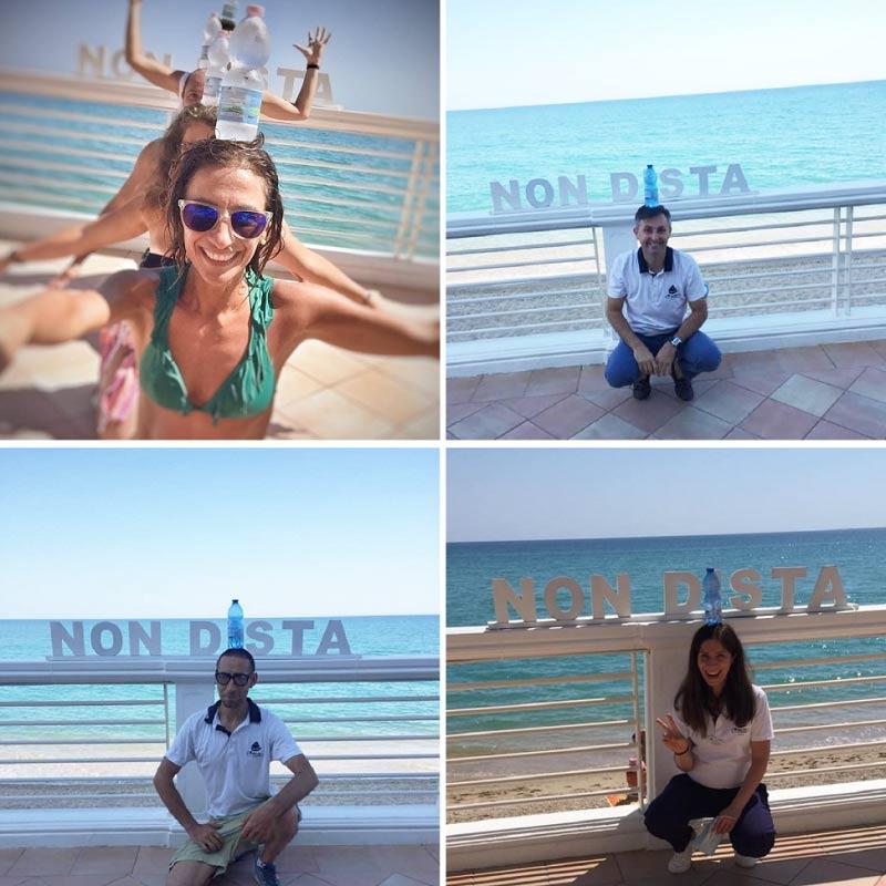 Persone posano con bottiglie d'acqua davanti alla scritta 'NON DISTA' sul mare.